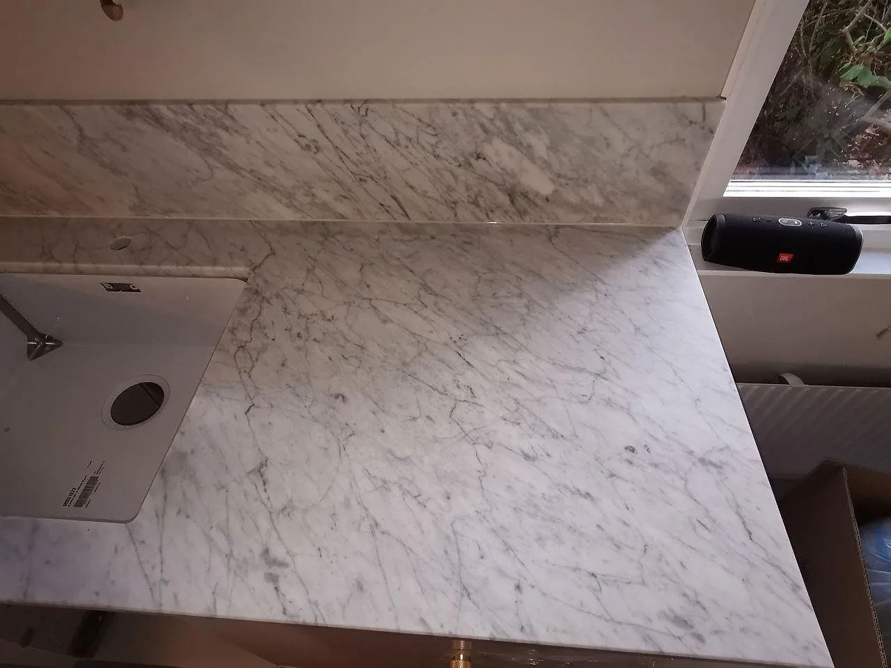 Närbild på matt Carrara CD-marmor vid fönstret med uppvikt stänkkant och underlimmad diskho i ett kök i Ramlösa.