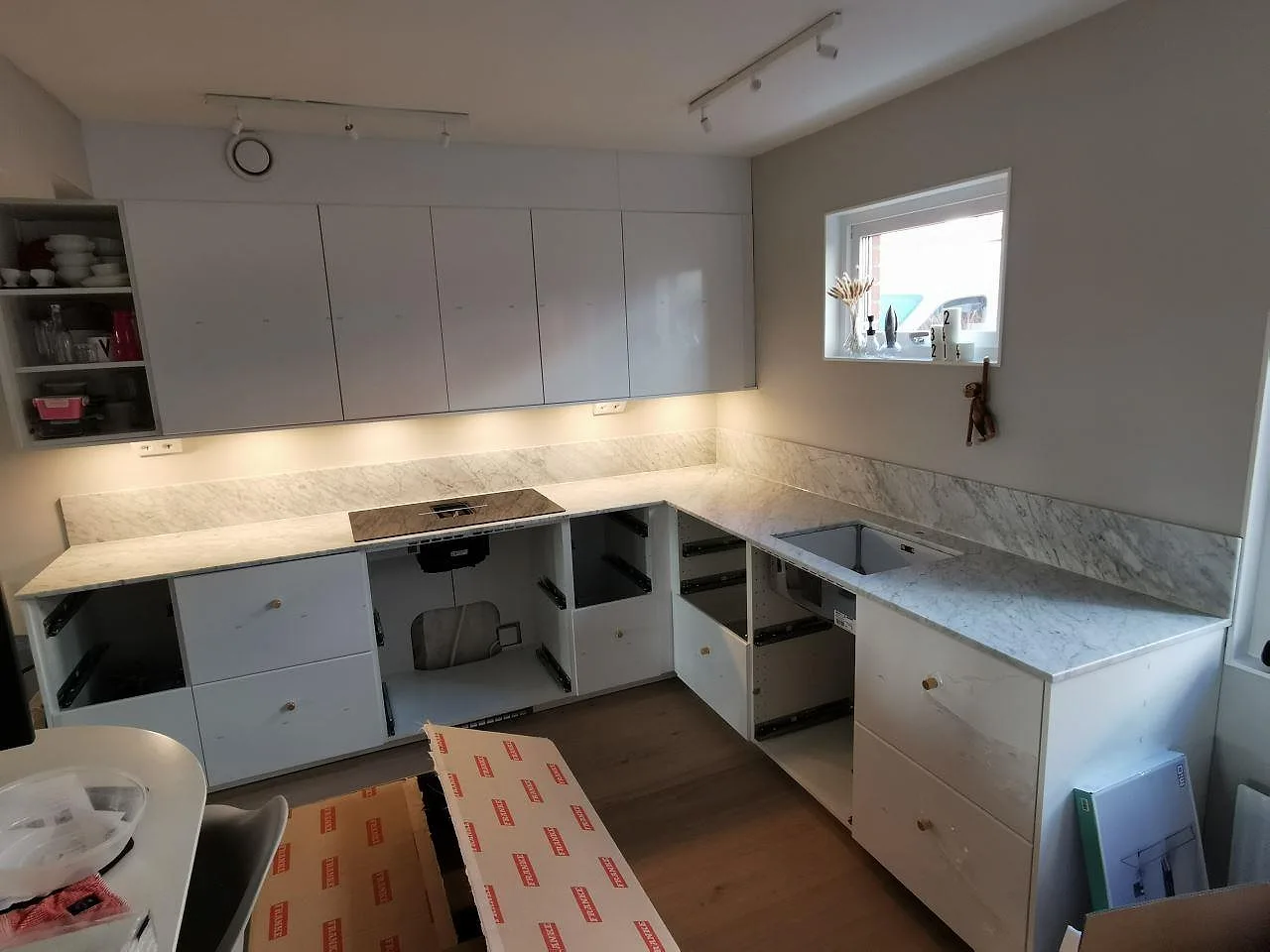 Köksbänkskiva i matt vit Carrara CD-marmor med grå ådring i ett vinkelställt kök i Ramlösa, med uppvikt stänkskydd, underlimmad diskho och utskärning för induktionshäll.