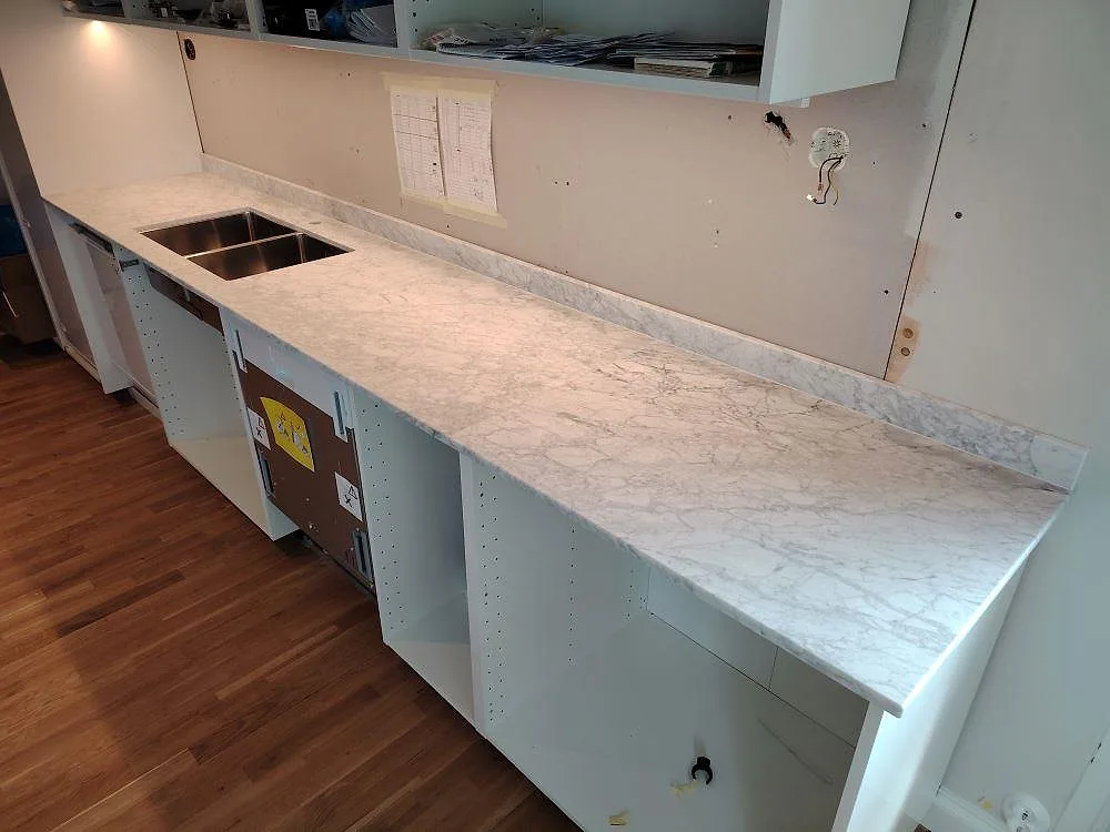 Köksbänkskiva i matt Carrara C marmor med låg stänkkant och synlig diskho under montering i ett kök i Saltsjö Boo.
