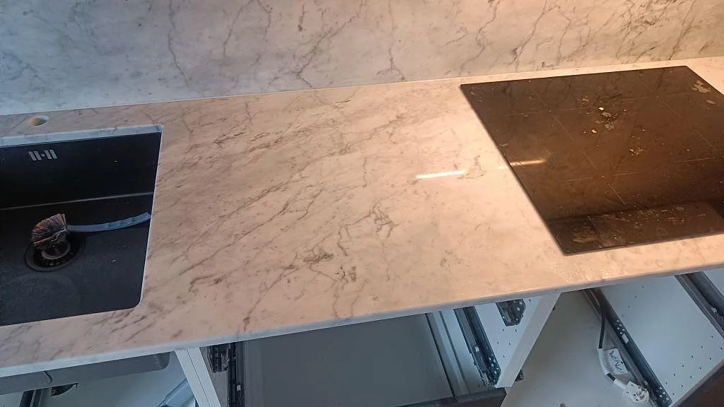 Detalj av polerad Carrara C marmorbänkskiva med svart diskho, induktionshäll och grå ådring i Skee.