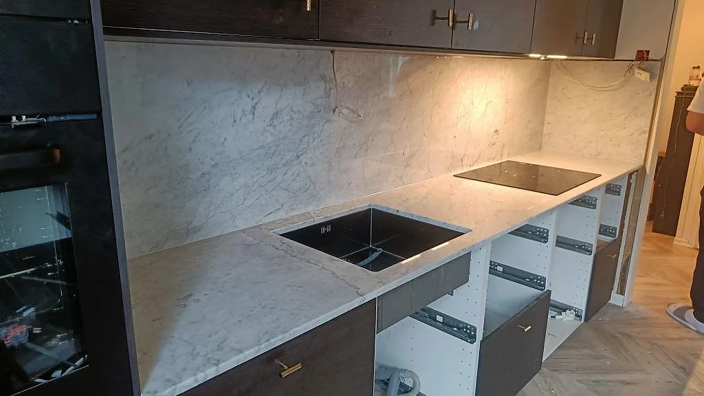 Köksbänkskiva i polerad Carrara C marmor med hög stänkvägg, underlimmad svart diskho och induktionshäll i ett kök i Skee.