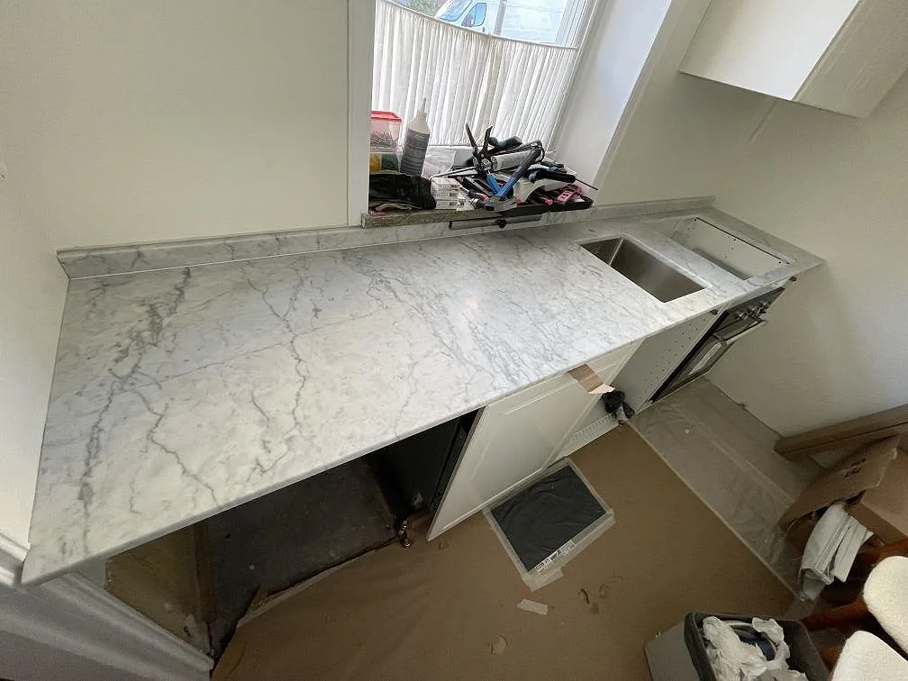 Köksbänkskiva i matt Carrara C marmor med grå ådring, uppvikt stänkkant och synlig diskhozon under ett fönster i Stockholm.