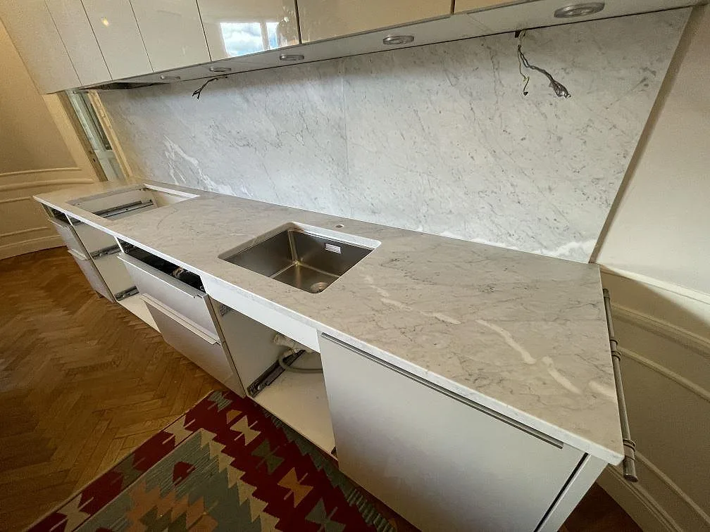 Detalj av matt Carrara C marmor vid diskhon, med ådrad bänkskiva och stenbeklädd vägg i ett kök i Stockholm.