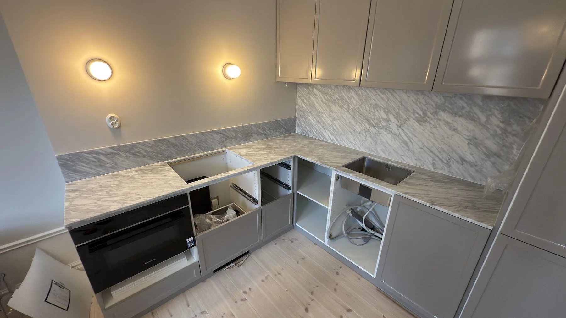 L-formad köksbänkskiva i matt Carrara C marmor, 20 mm, med marmorstänkskydd och grå skåp under montering i Stockholm.