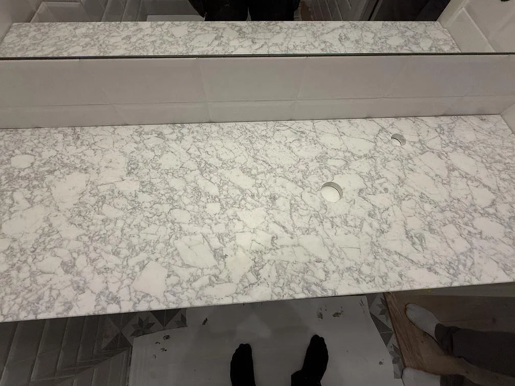 Detalj av badrumsbänkskiva i matt Carrara C marmor i Stockholm, med tät grå ådring och spegelkant bakom.