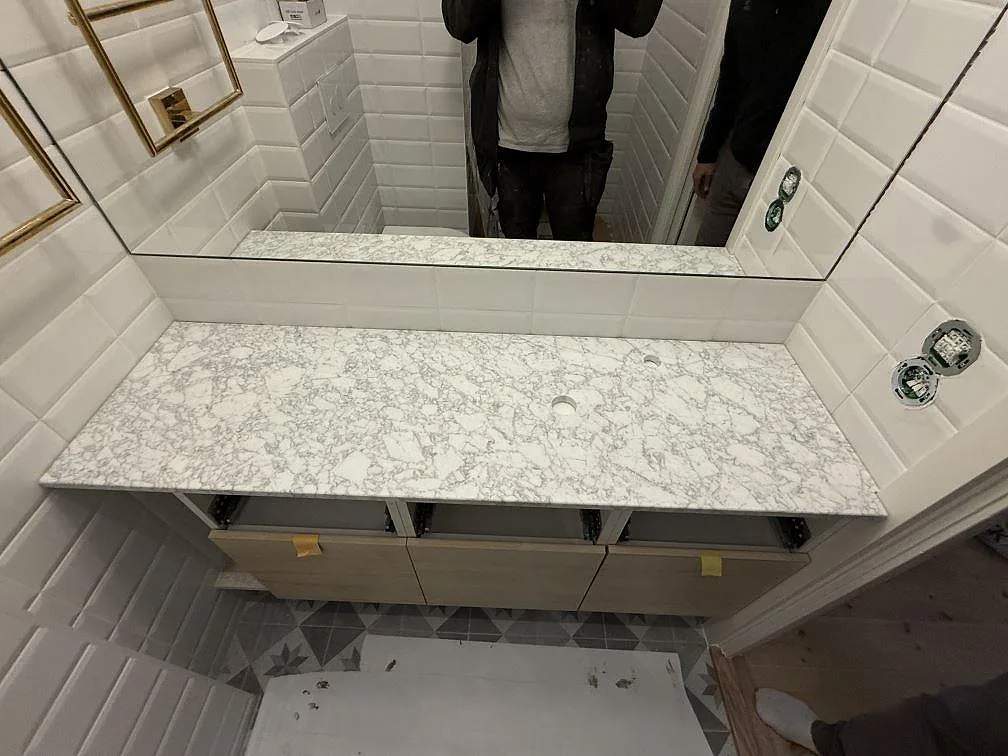 Badrumsbänkskiva i matt Carrara C marmor, 20 mm, monterad framför spegel och kakel i Stockholm.