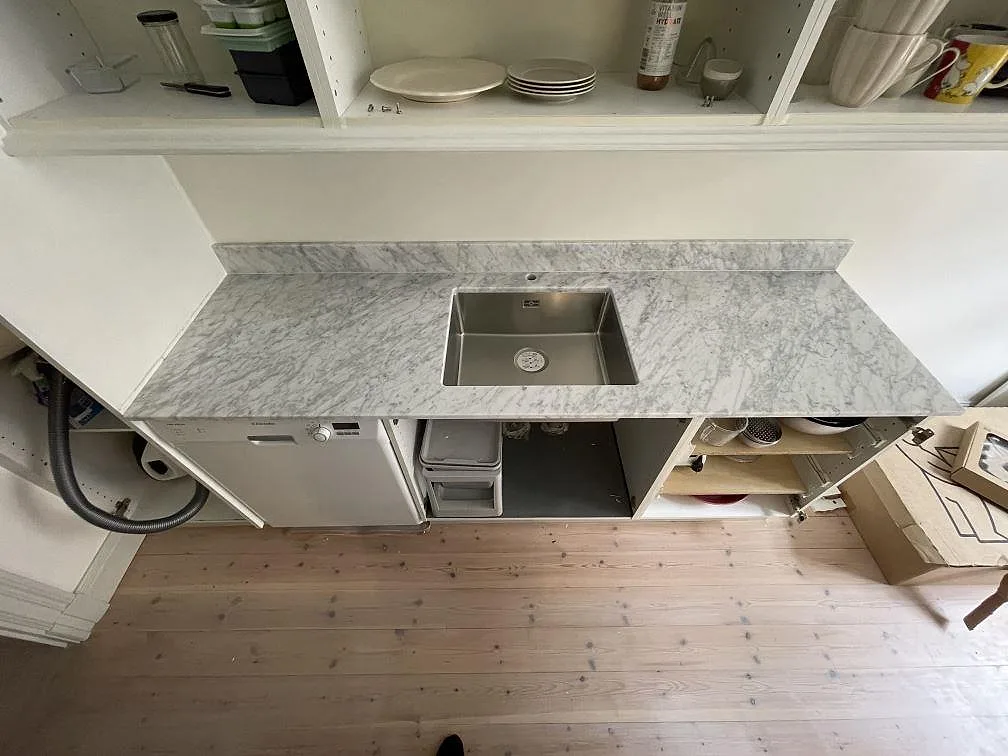 Underlimmad fyrkantig diskho i rostfritt stål i en köksbänkskiva av Carrara CD-marmor med uppvikt stänkkant i ett kök i Stockholm.