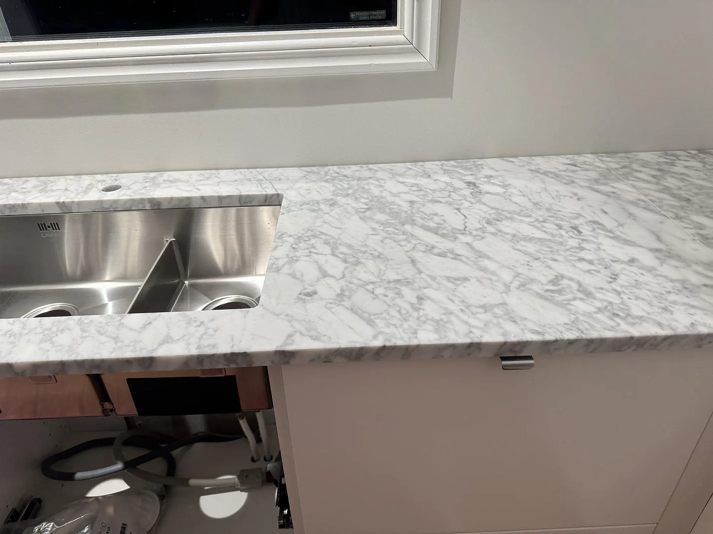 Köksbänkskiva i matt Carrara C marmor med rostfri dubbelho under fönster i ett kök i Stora höga.