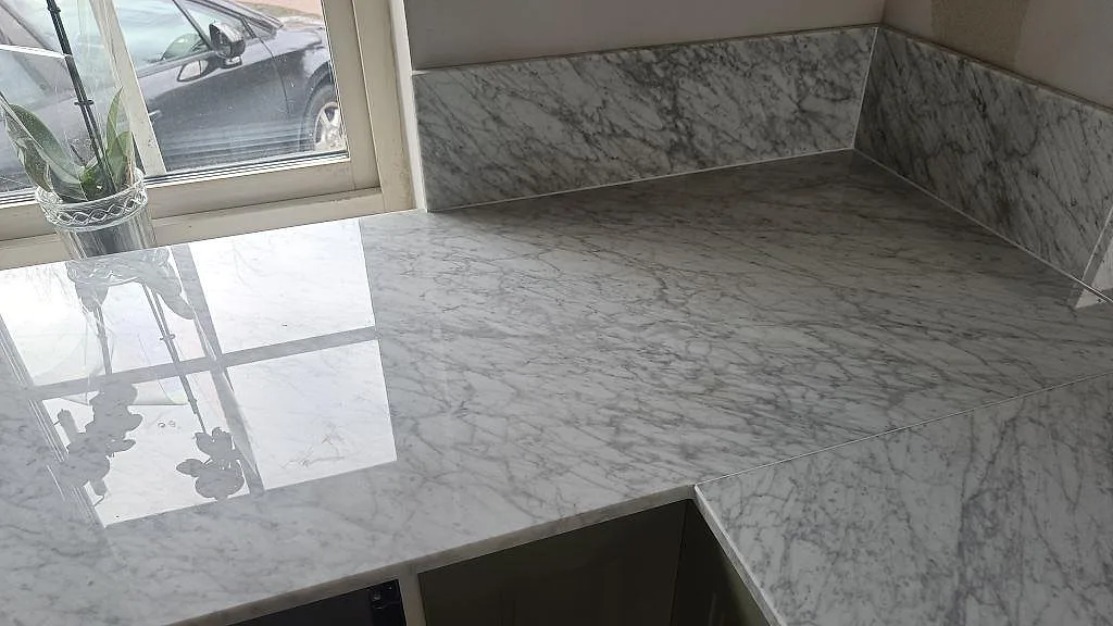 Polerad Carrara C marmorbänkskiva vid fönster med vas, uppvikt stänkkant och grön köksinredning i Svanshall.