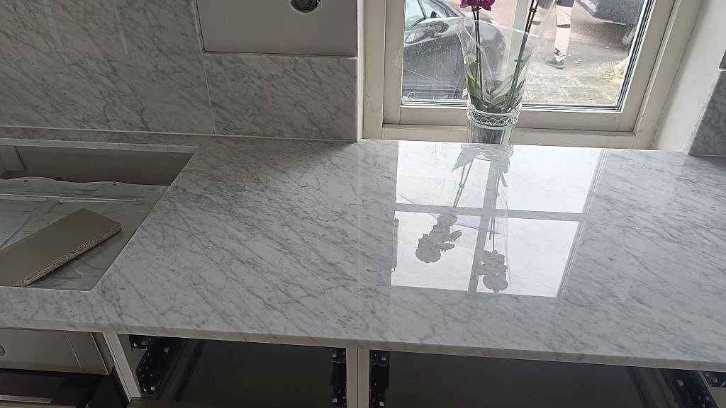 Carrara C köksbänkskiva med blank reflektion från fönster och vas, monterad över gröna underskåp i Svanshall.