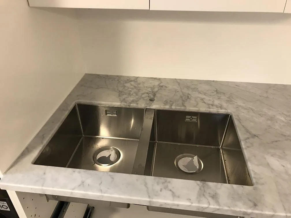Köksbänkskiva i vit Carrara marmor med grå ådring, matt yta, med två underlimmade rostfria diskhoar i ett kök i Trelleborg.