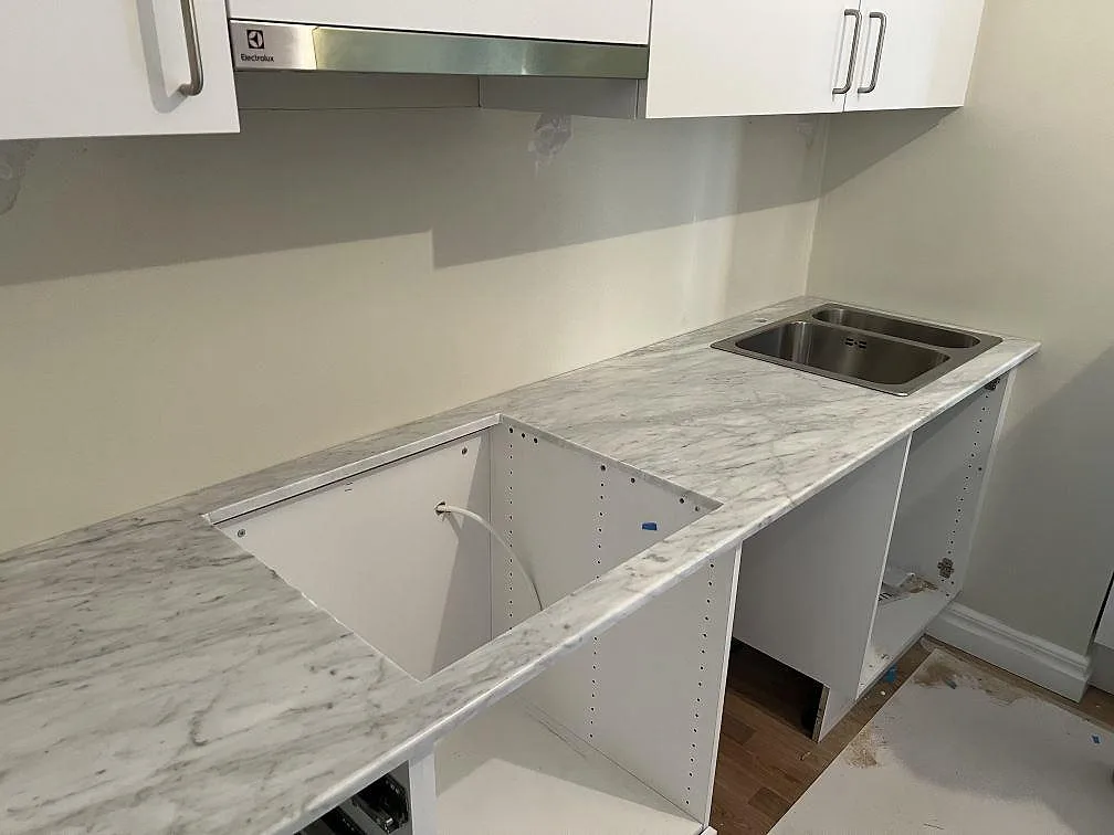 Köksbänkskiva i 20 mm polerad Carrara C marmor med övermonterad rostfri dubbelho, öppet hällurtag, vita skåp och fläkt i ett kök i Trollhättan.