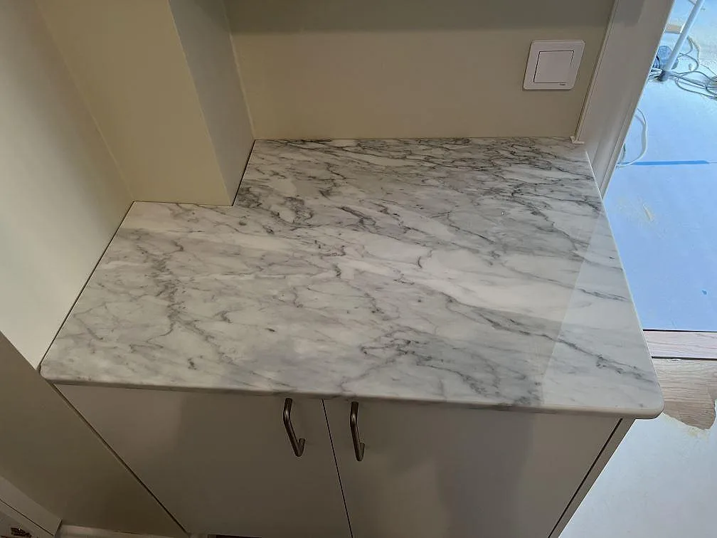 Sidobänk i polerad Carrara C marmor monterad på vita luckor vid en ljus vägg i köket i Trollhättan.