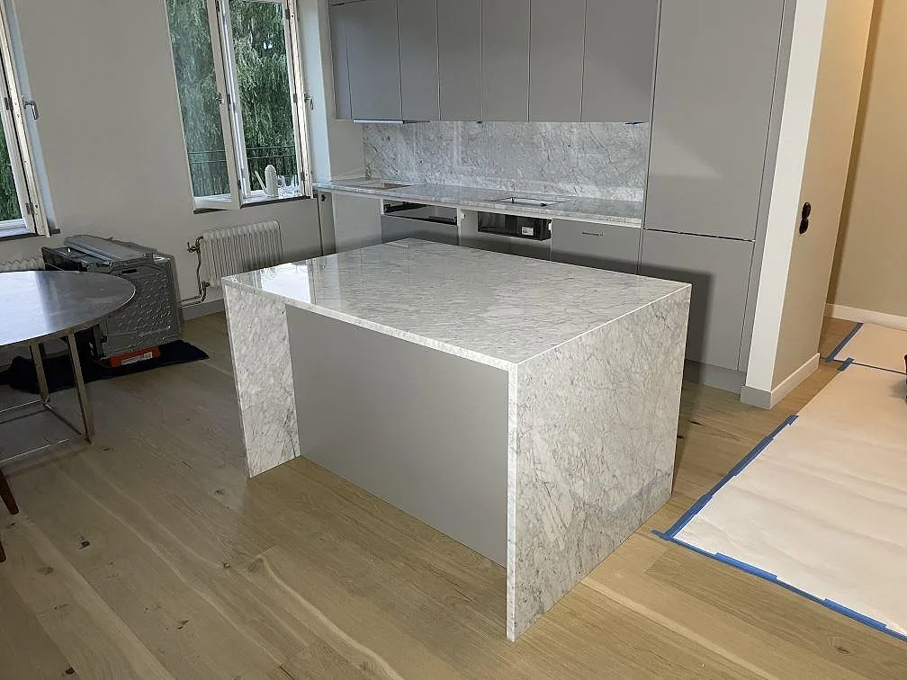 Polerad köksbänkskiva i Carrara C marmor med stor köksö, stenklädda sidor och stänkskydd i ett kök i Uppsala.