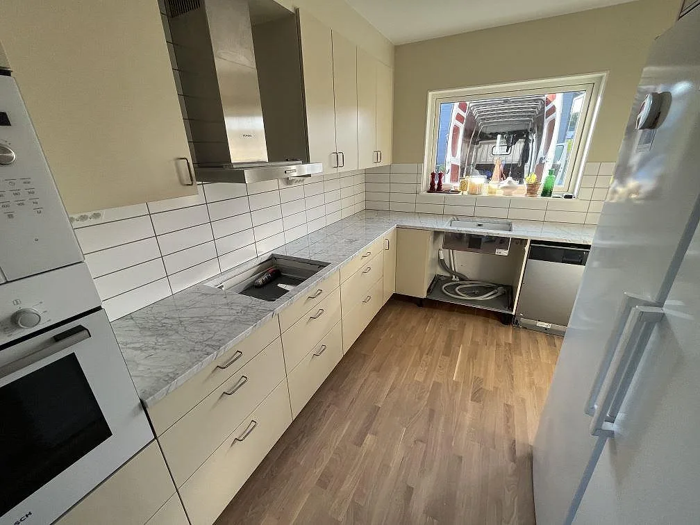 Köksbänkskiva i polerad Carrara C marmor i ett L-format kök i Västerås med häll, vit kakelvägg och fläktkåpa.