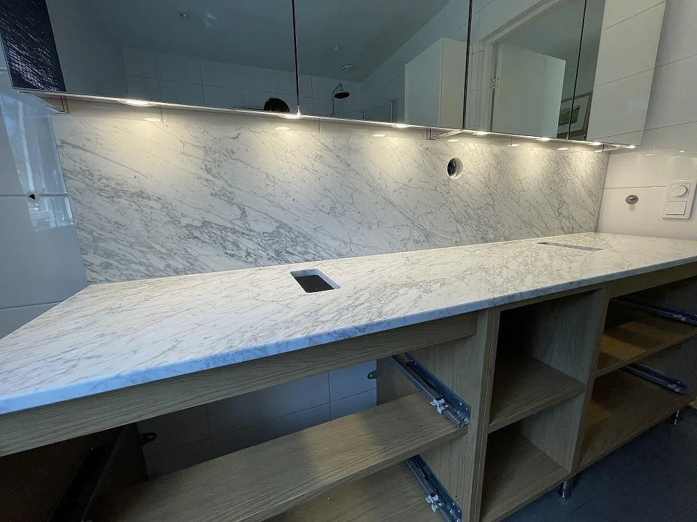 Badrumsbänkskiva i matt Carrara C marmor, 20 mm, med matchande väggbeklädnad och rektangulära urtag under montering i Vaxholm.