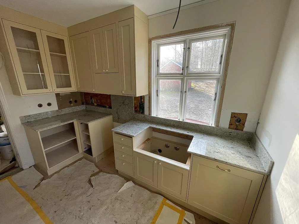 Köksbänkskiva i polerad Colonial White-granit, ljusgrå med svart spräcklighet, monterad i L-form runt ett fönster i ett kök i Östra Sönnarslöv.