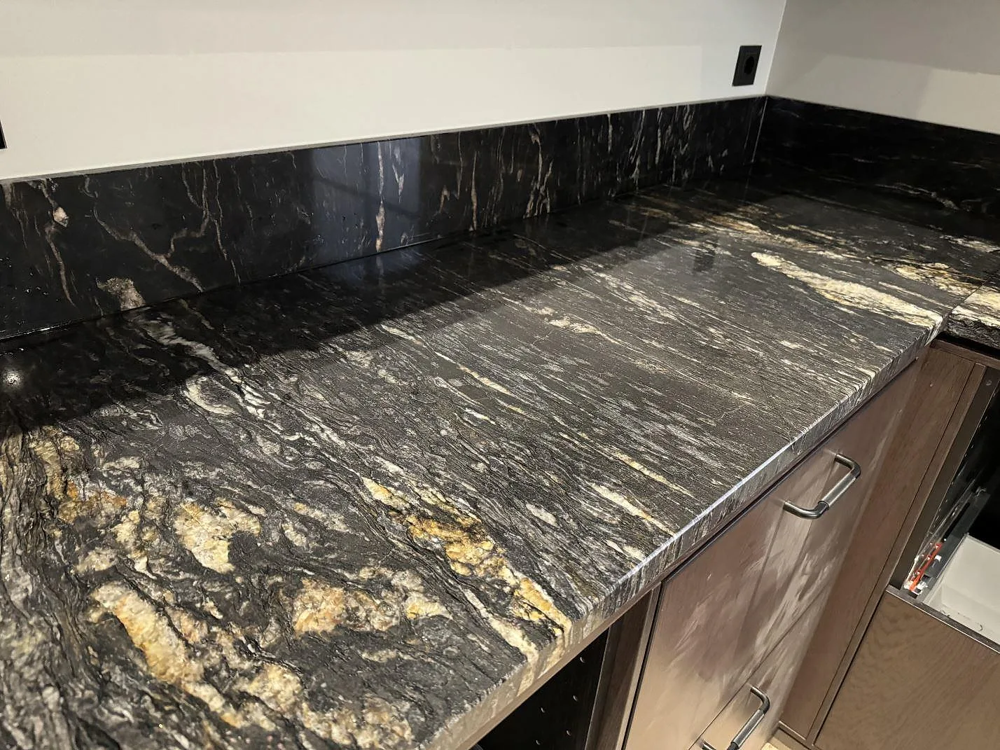 Längsgående köksbänkskiva i polerad Cosmic Black granit med uppvikt stänkskydd och träinredning i Grebbestad.