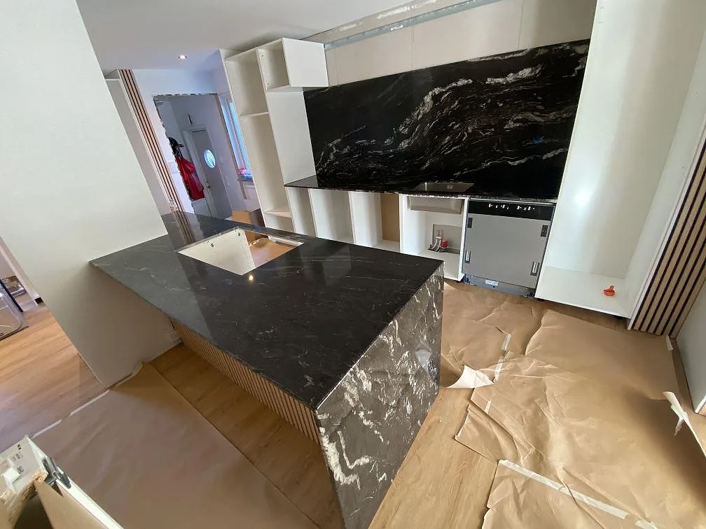 Köksbänkskiva i svart Cosmic Black granit, polerad, med köksö och heltäckande stänkvägg i samma sten i ett kök under montering i Kävlinge.