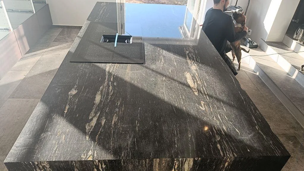 Köksö i Cosmic Black-granit med nedfälld fläkt i hällen och dramatisk ådring, med havsutsikt genom fönstret i ett kök i Laholm.