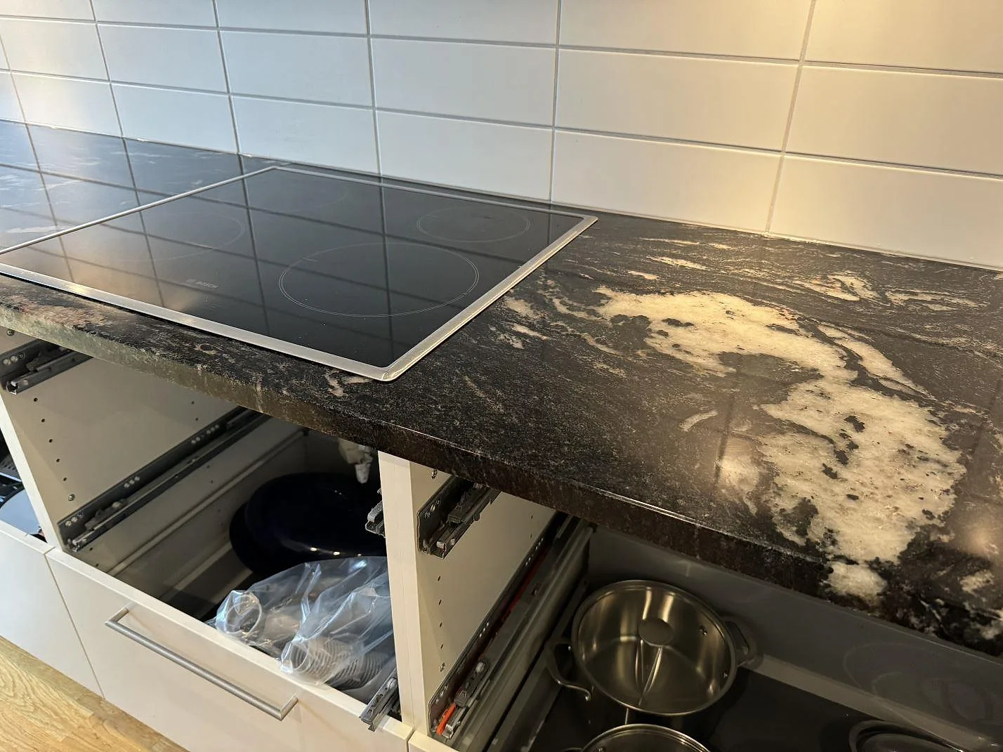 Köksbänkskiva i polerad Cosmic Black granit med induktionshäll och öppna vita skåpstommar i Malmö.