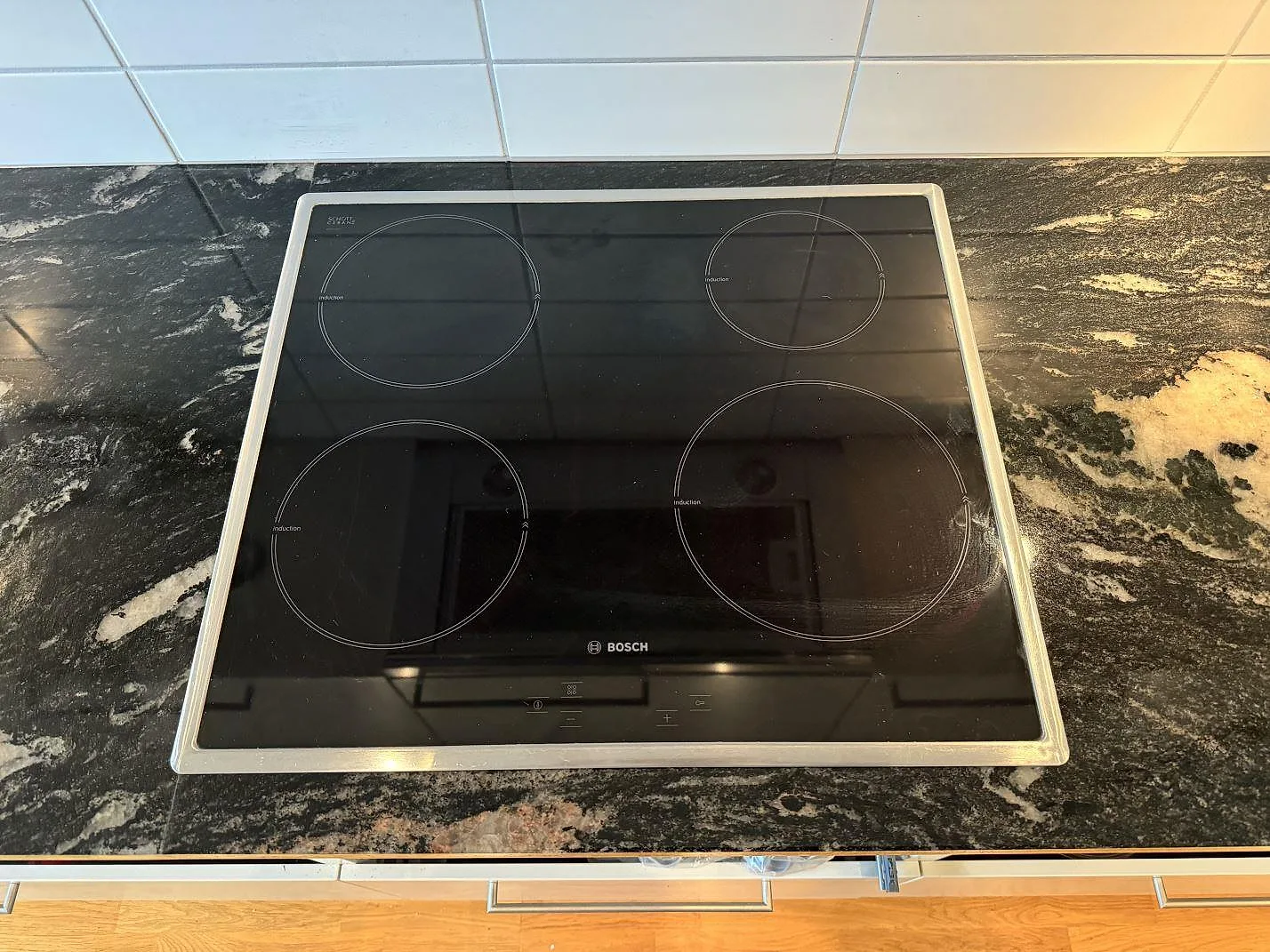 Induktionshäll infälld i polerad köksbänkskiva av Cosmic Black granit framför vitt kakel i Malmö.