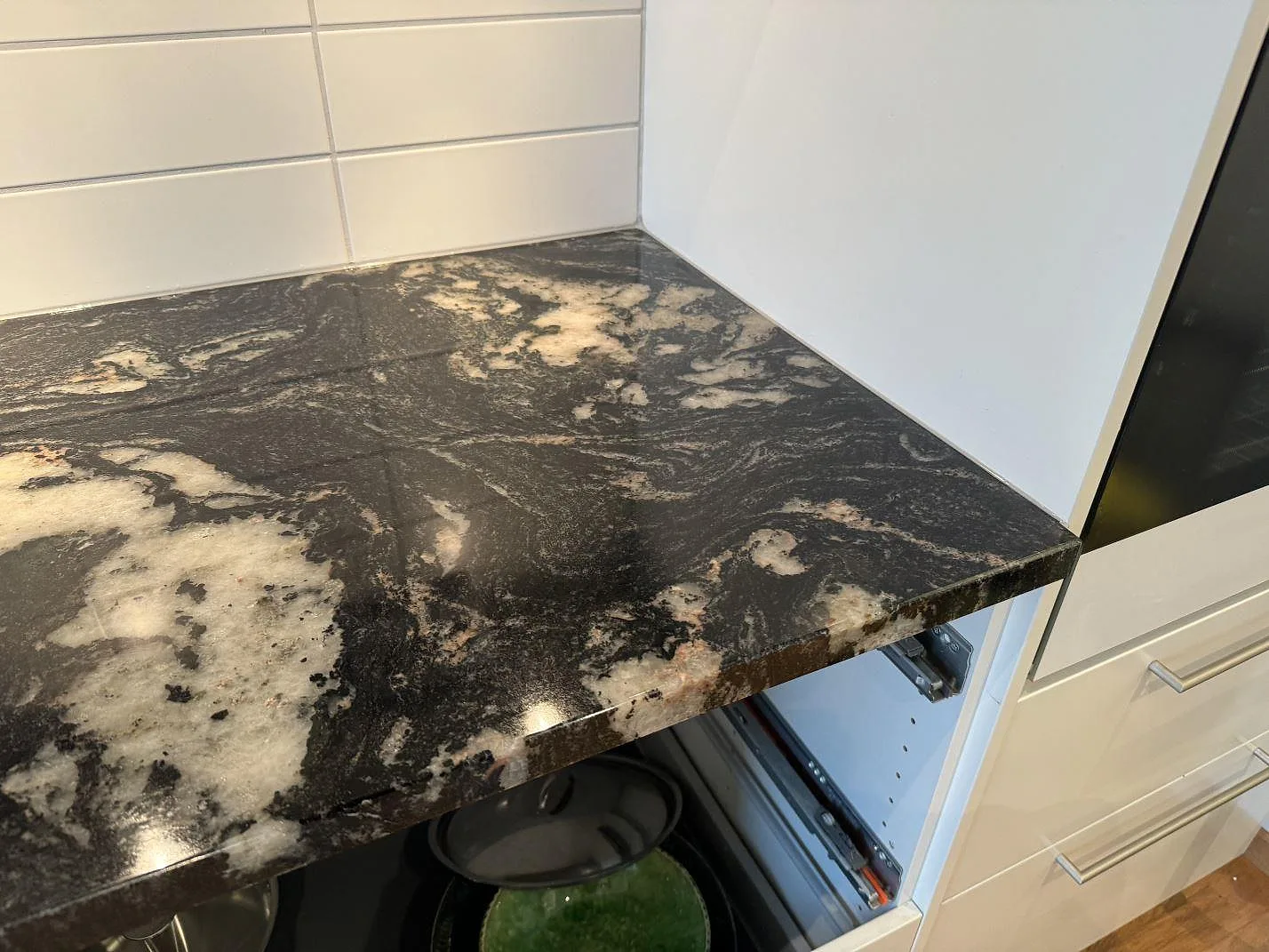 Närbild på polerad köksbänkskiva i Cosmic Black granit med vit kakelvägg och öppen skåpstomme i Malmö.