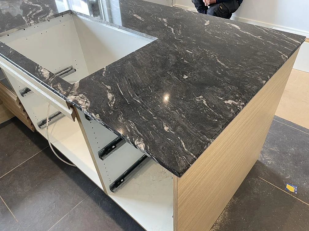 Köksö i Cosmic Black granit med polerad yta, vit stomme och träpanel under montering i Tølløse.