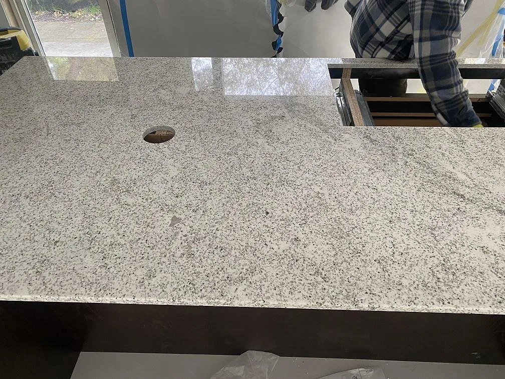 Köksbänkskiva i polerad Cotton White granit med hällurtag och rund genomföring under installation i Höllviken.