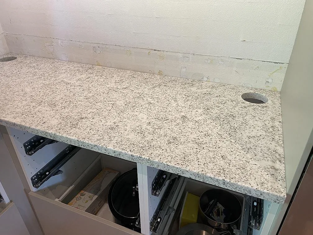 Lång köksbänkskiva i Cotton White granit med två runda håltagningar och öppna stommar under montering i Höllviken.