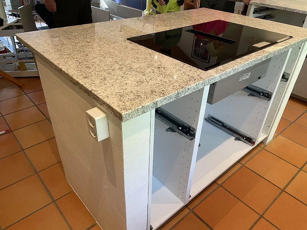 Köksö i ljus Cotton White granit med svart induktionshäll, öppna vita stommar och terrakottafärgat klinkergolv i Höllviken.