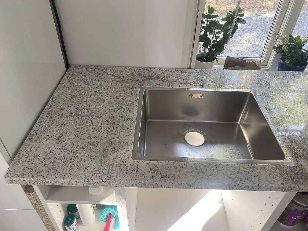 Närbild på underlimmad rostfri diskho i polerad Cotton White-granit med tydliga gråaktiga korn i Vålberg.