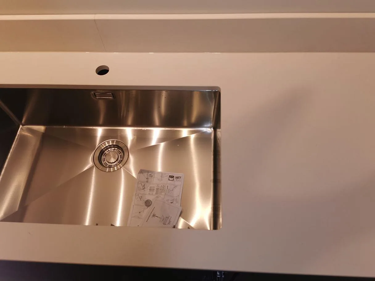 Underlimmad rostfri diskho i matt vit Dekton Aeris köksbänkskiva med blandarehål i ett kök i Lund.