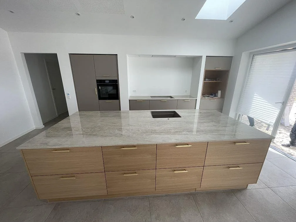 Köksbänkskiva i Dekton Arga, polerad gråbeige keramik, på en stor köksö med ekfronter och mässingshandtag i ett nybyggt kök i Agedrup.