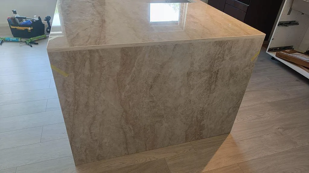 Vattenfallssida på köksö i polerad Dekton Arga keramik med beige ådring och öppna stommar under montering i Alvesta.
