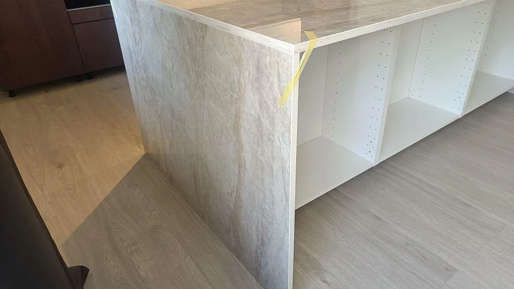 Sidovy av köksö med vattenfallssida i polerad Dekton Arga och vita öppna köksstommar under montering i Alvesta.