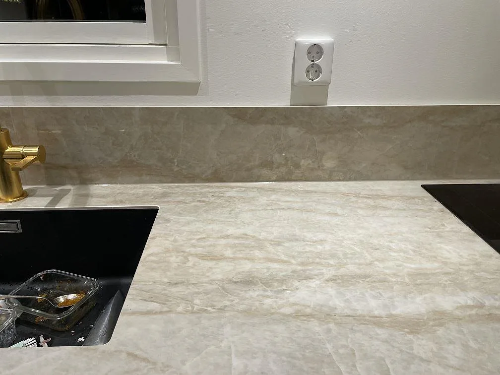 Ljus köksbänkskiva i polerad Dekton Arga med uppvikt stänkskydd, svart diskho och vägguttag i Billdal.