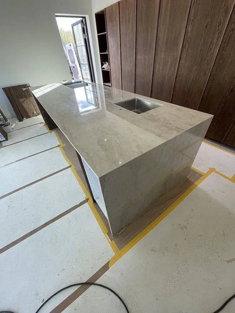 Köksö i polerad Dekton Arga, ljus gråbeige keramik med marmorlikt mönster, med urtag för diskho i ett kök under montering i Borlänge.