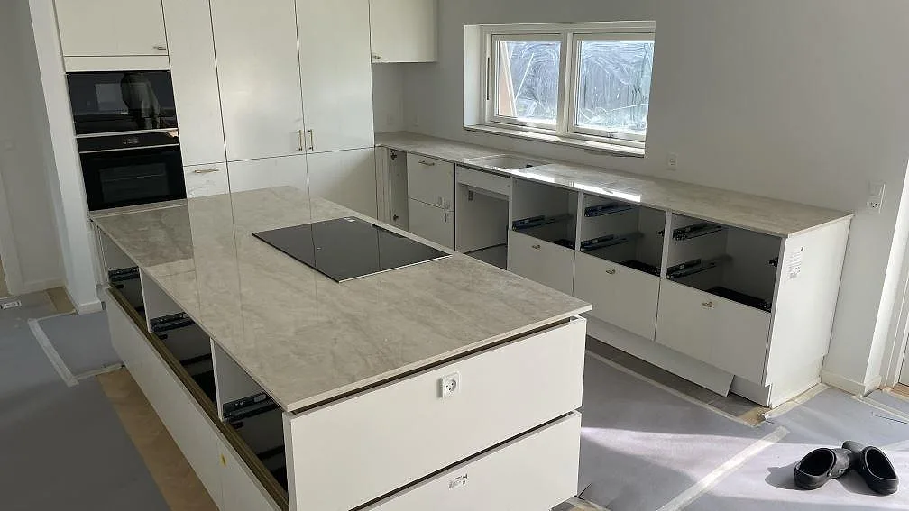 Köksbänkskiva i polerad Dekton Arga, keramik, med köksö och bänkrad under fönster i ett kök under montering i Brøndby Strand.