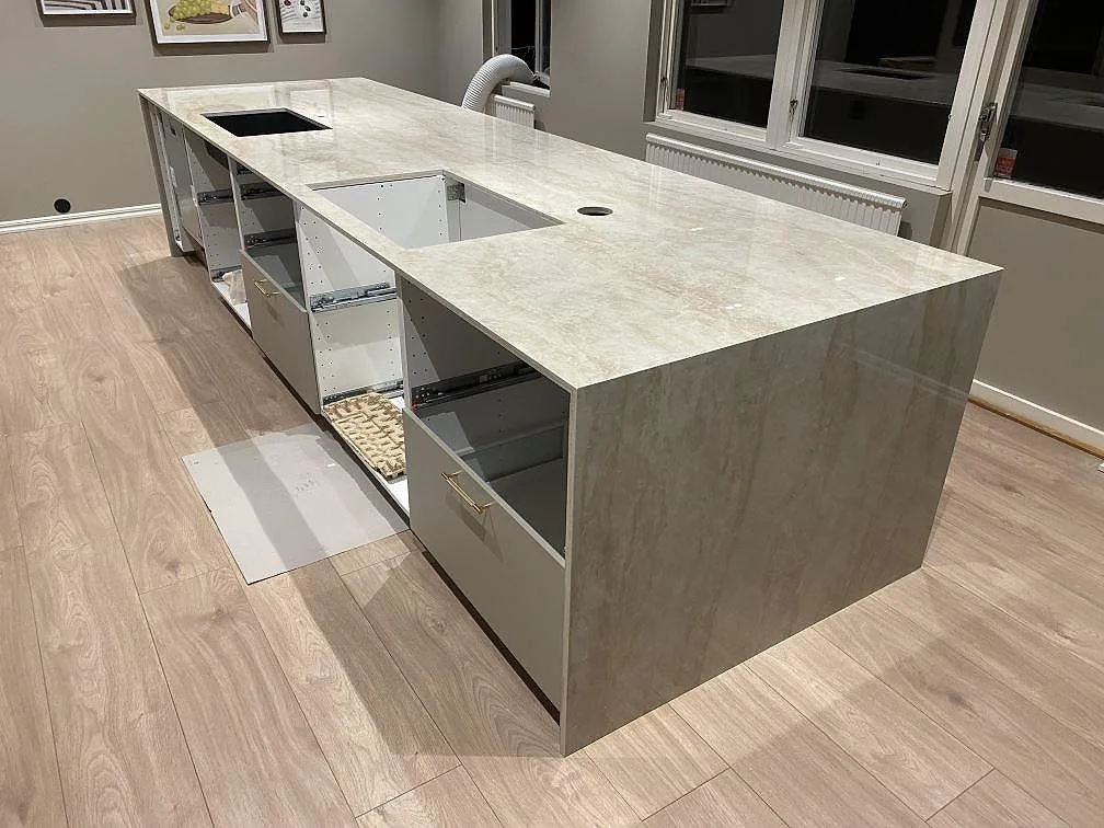 Polerad Dekton Arga bänkskiva med stenklädd gavel, runt blandarhål och öppna stommar i ett kök under montering i Fjärås.