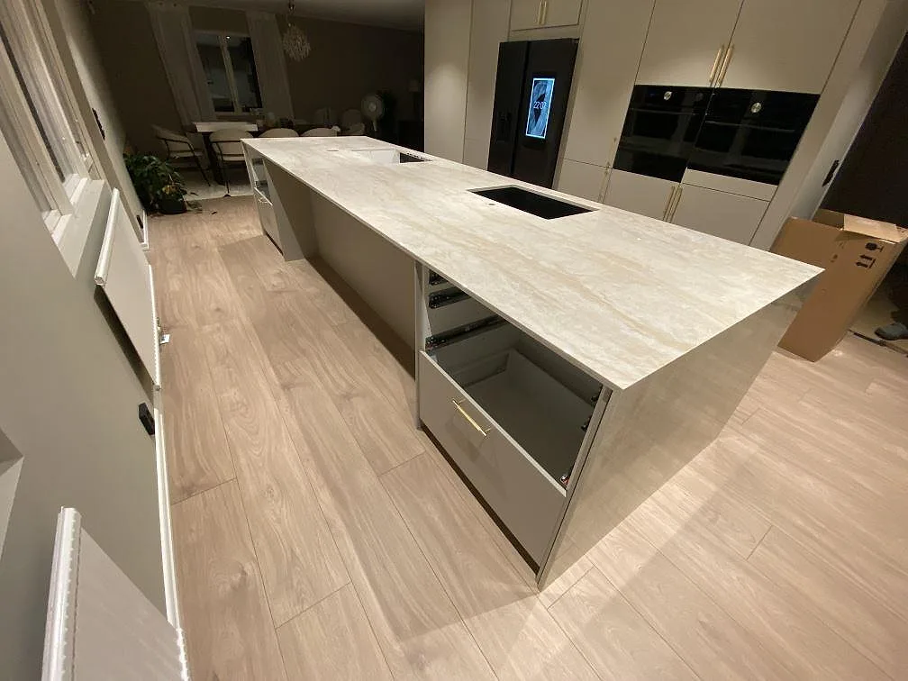 Lång köksö med köksbänkskiva i polerad Dekton Arga, svart diskho och hällurtag framför köksskåp i Fjärås.