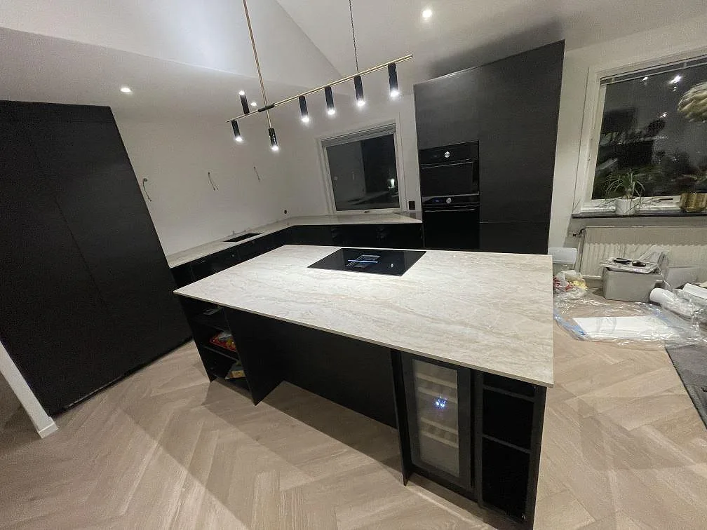 Köksö i polerad Dekton Arga keramik med infälld induktionshäll i ett kök under montering i Frillesås, med parkettgolv och svarta köksluckor.