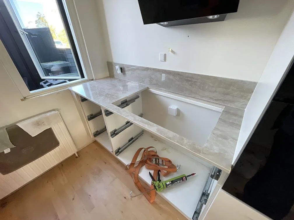 L-formad köksbänkskiva i polerad Dekton Arga keramik med hälluttag, stänkskydd och öppna vita köksstommar i Glostrup.