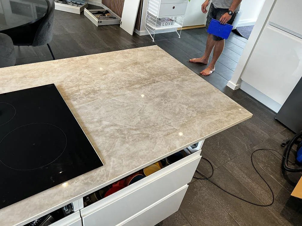 Polerad Dekton Arga på köksö med svart häll, öppna skåpsstommar och mörkt golv under montering i Hörby.