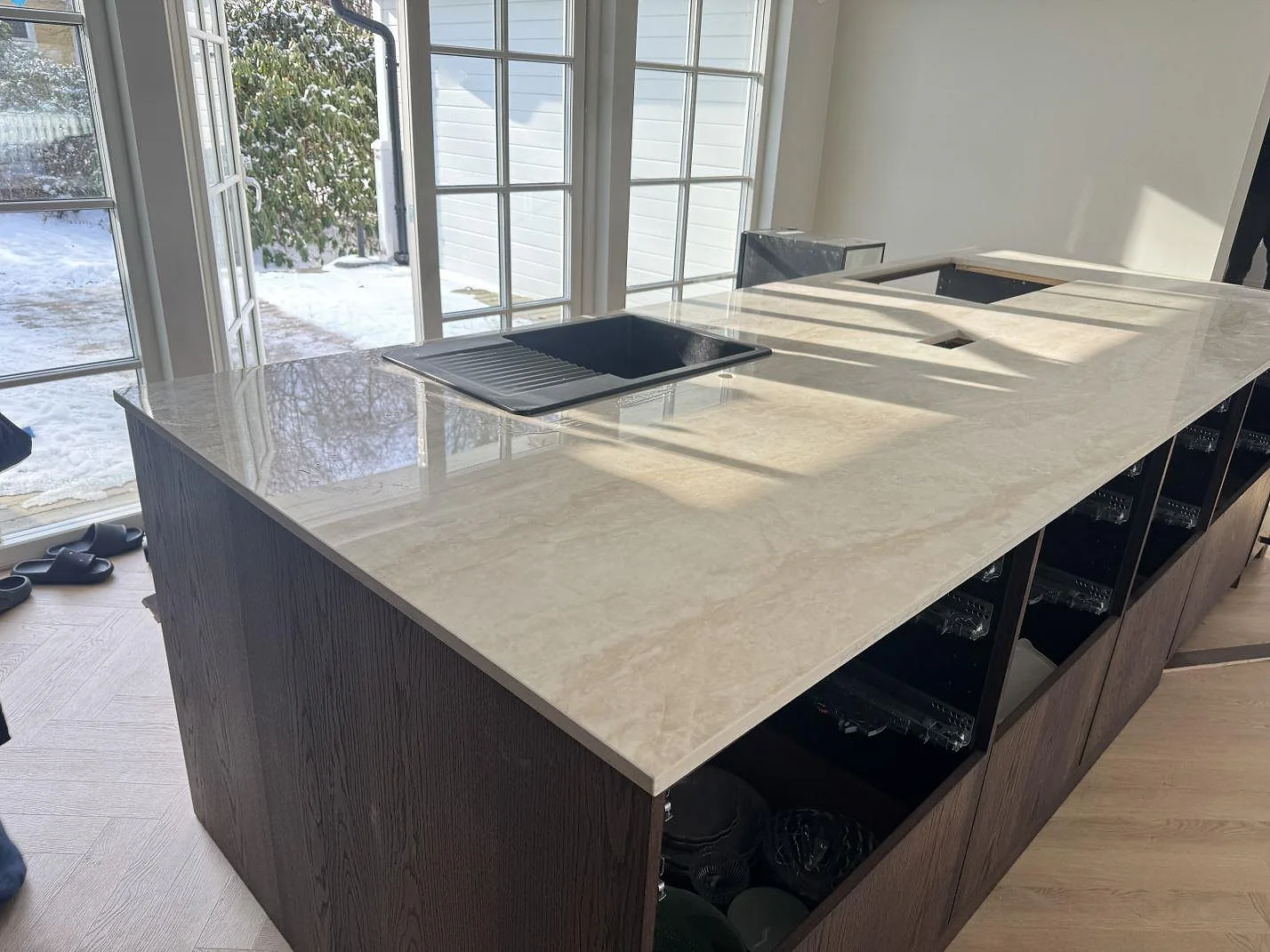 Köksbänkskiva och stor köksö i polerad Dekton Arga, ljust krämbeige keramik, med underlimmad svart komposithoar vid altandörrar i ett kök i Hörby.