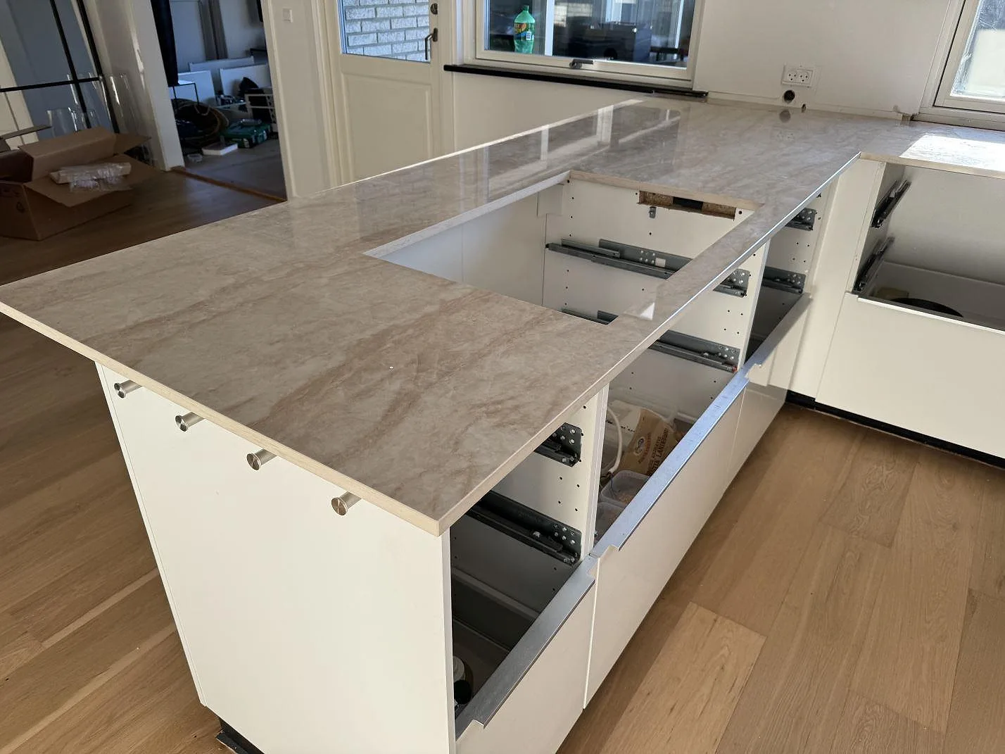 Köksö i polerad Dekton Arga med travertinlik ådring, vita köksstommar och öppna lådor under montering i Hørsholm.