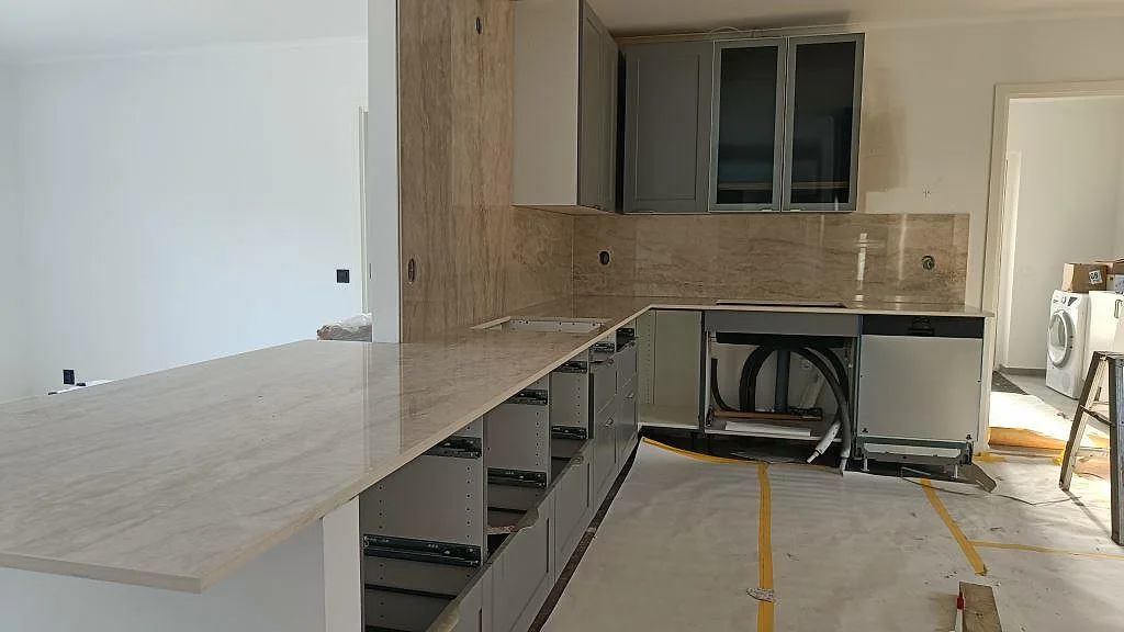 Köksbänkskiva och köksö i polerad beige Dekton Arga i ett kök i Kärna, fotograferat under montering med öppna stommar och heltäckande stänkvägg i samma sten.