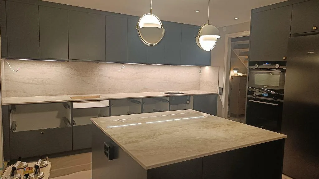 Köksbänkskiva i ljust grå Dekton Arga, polerad, med heltäckande stänkvägg i samma sten och en stor köksö i ett kök i Kalmar.