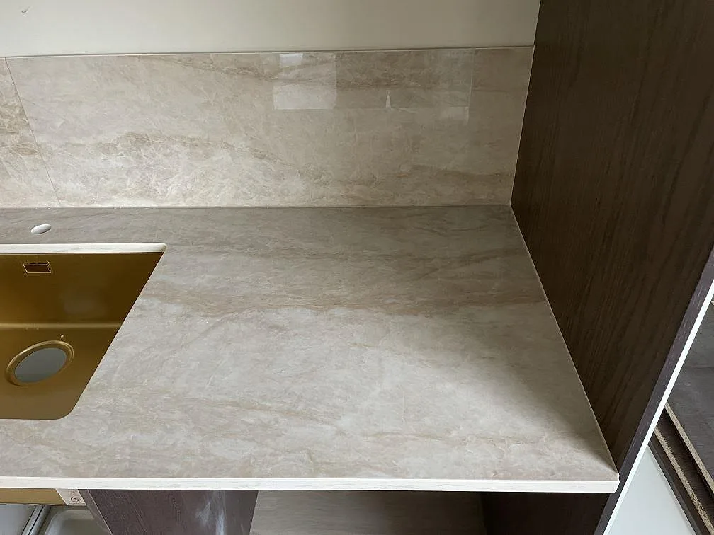Köksbänkskiva i Dekton Arga, polerad keramik i varm beige med mjuk ådring, med uppvikt stänkskydd och underlimmad guldfärgad diskho i ett kök i Karlshamn.