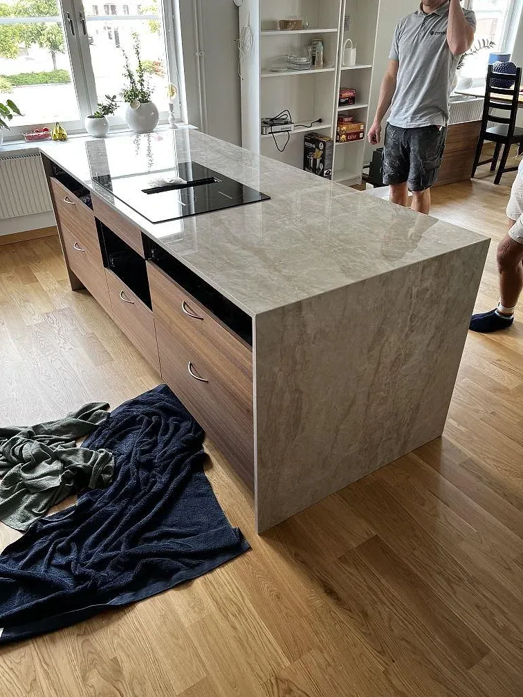 Sidovy av köksö i polerad Dekton Arga med häll, träinredning och ekgolv under montering i Karlskrona.